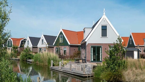 Une rangée de maisons avec un quai s’avançant dans une étendue d’eau calme, le tout entouré de verdure.