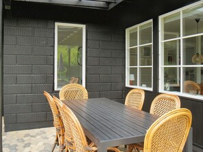 Un patio con una mesa de madera oscura y sillas de mimbre, rodeado de muros de ladrillo negro y ventanas amplias.