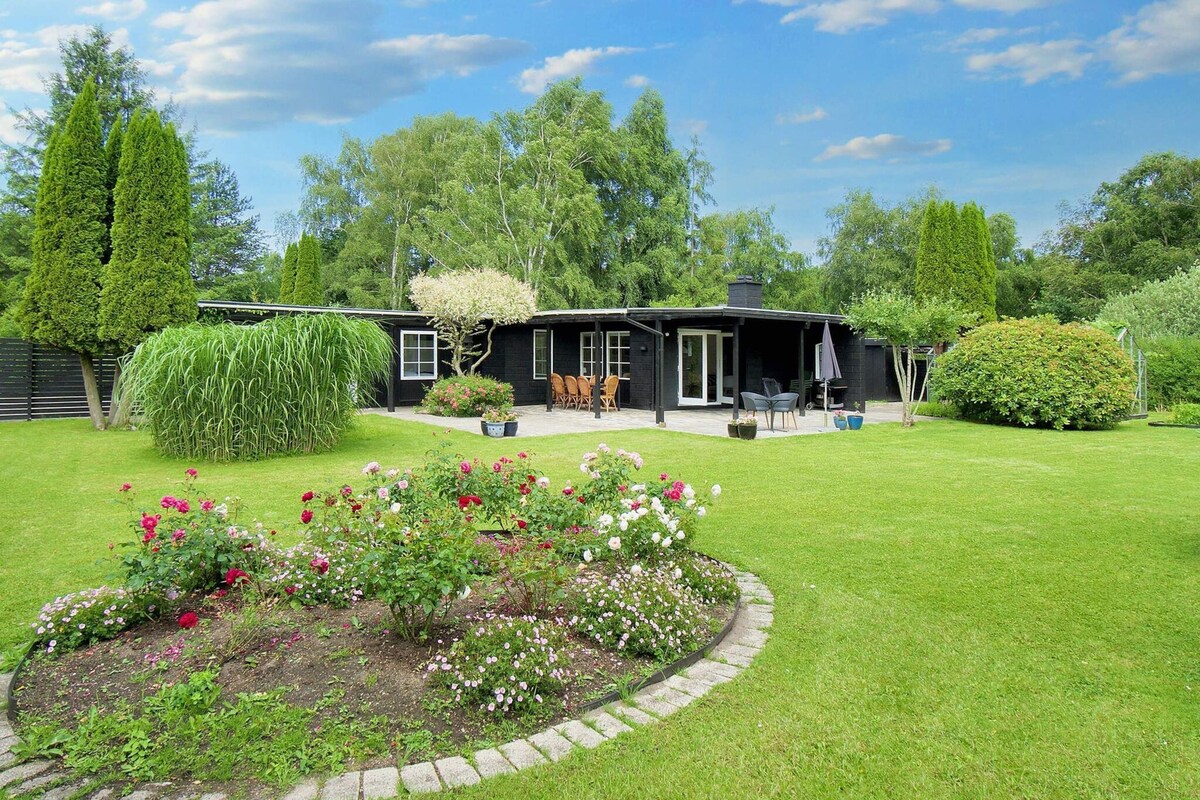 Une maison noire dotée d’un patio, entourée d’un jardin bien entretenu où poussent diverses plantes et fleurs.
