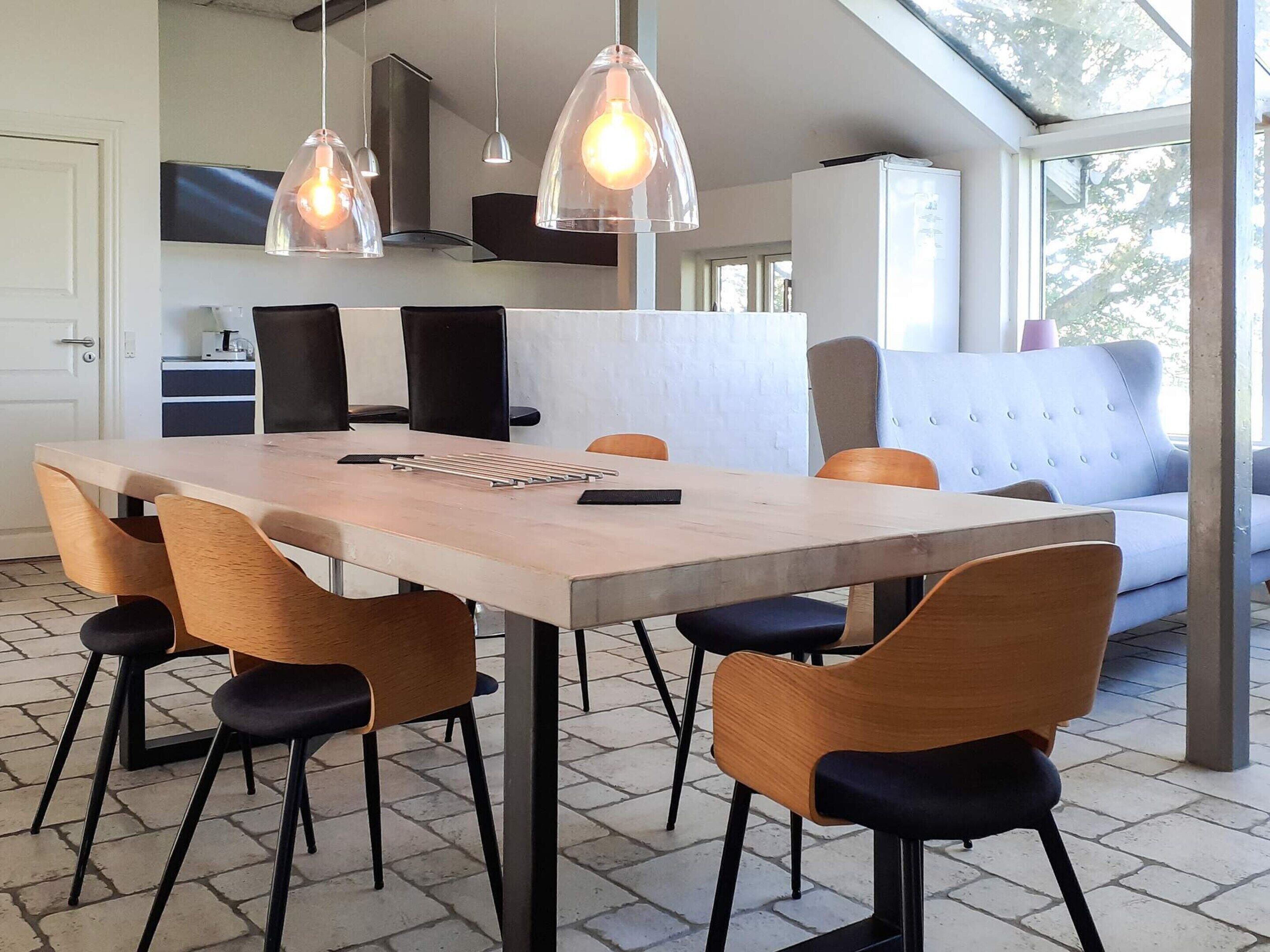 A modern kitchen with a dining area, featuring a large table, chairs, pendant lights, and a sofa.