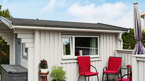 Ein kleines weißes Haus mit einer Terrasse, zwei roten Stühlen, einer grauen Aufbewahrungsbox und einem Blumentopf.