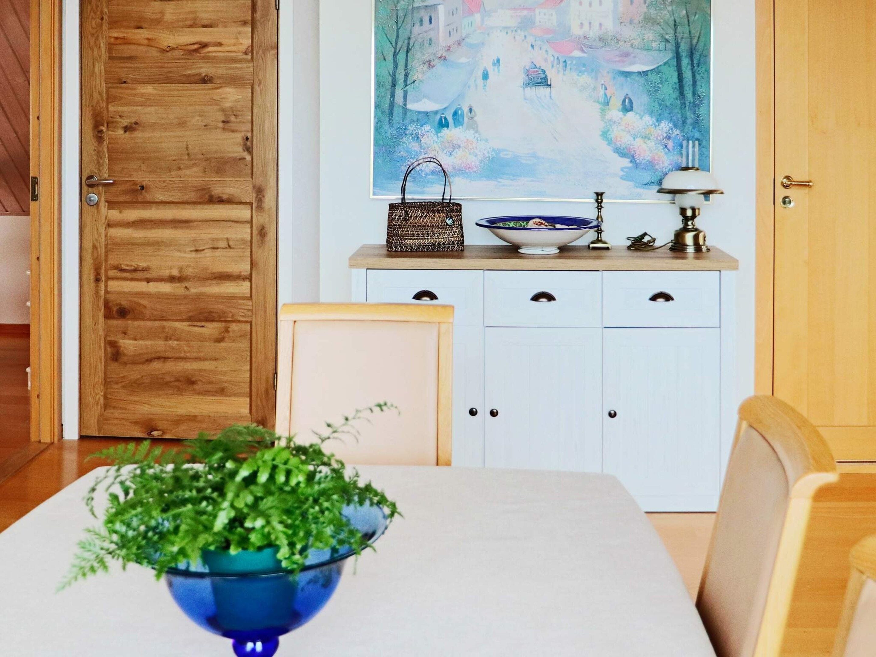 A dining area with a table, chairs, a blue bowl with green plants, a white cabinet with drawers, and a painting on the wall.