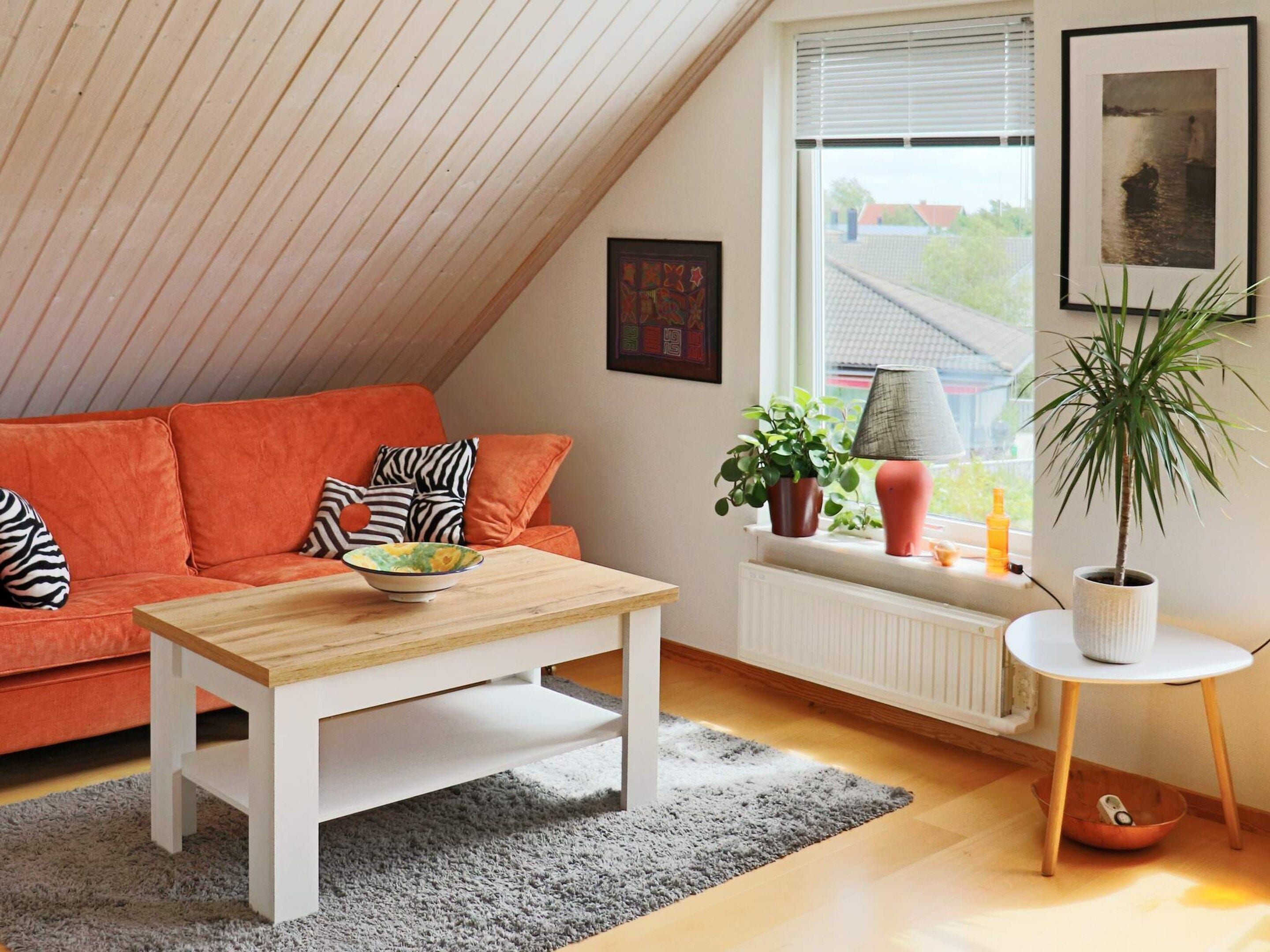 A cozy living room with a sloped ceiling, a wooden coffee table, a red sofa with patterned cushions, a grey rug, a white radiator, and a window with blinds.