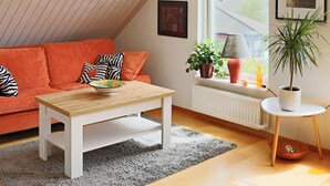 Un salon chaleureux avec un plafond en pente, une table basse en bois, un canapé rouge agrémenté de coussins à motifs, un tapis gris, un radiateur blanc et une fenêtre avec des stores.