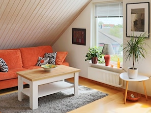 A cozy living room with a sloped ceiling, a wooden coffee table, a red sofa with patterned cushions, a grey rug, a white radiator, and a window with blinds. - 4 Star Holiday Home in Frillesas (Frillesas)