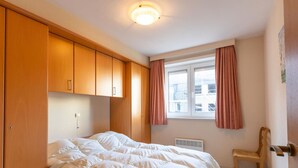A bedroom with a bed, wooden wardrobe, a window with curtains, a radiator, and a chair. - Apartment in Middelkerke Near Sandy Beach (Middelkerke)