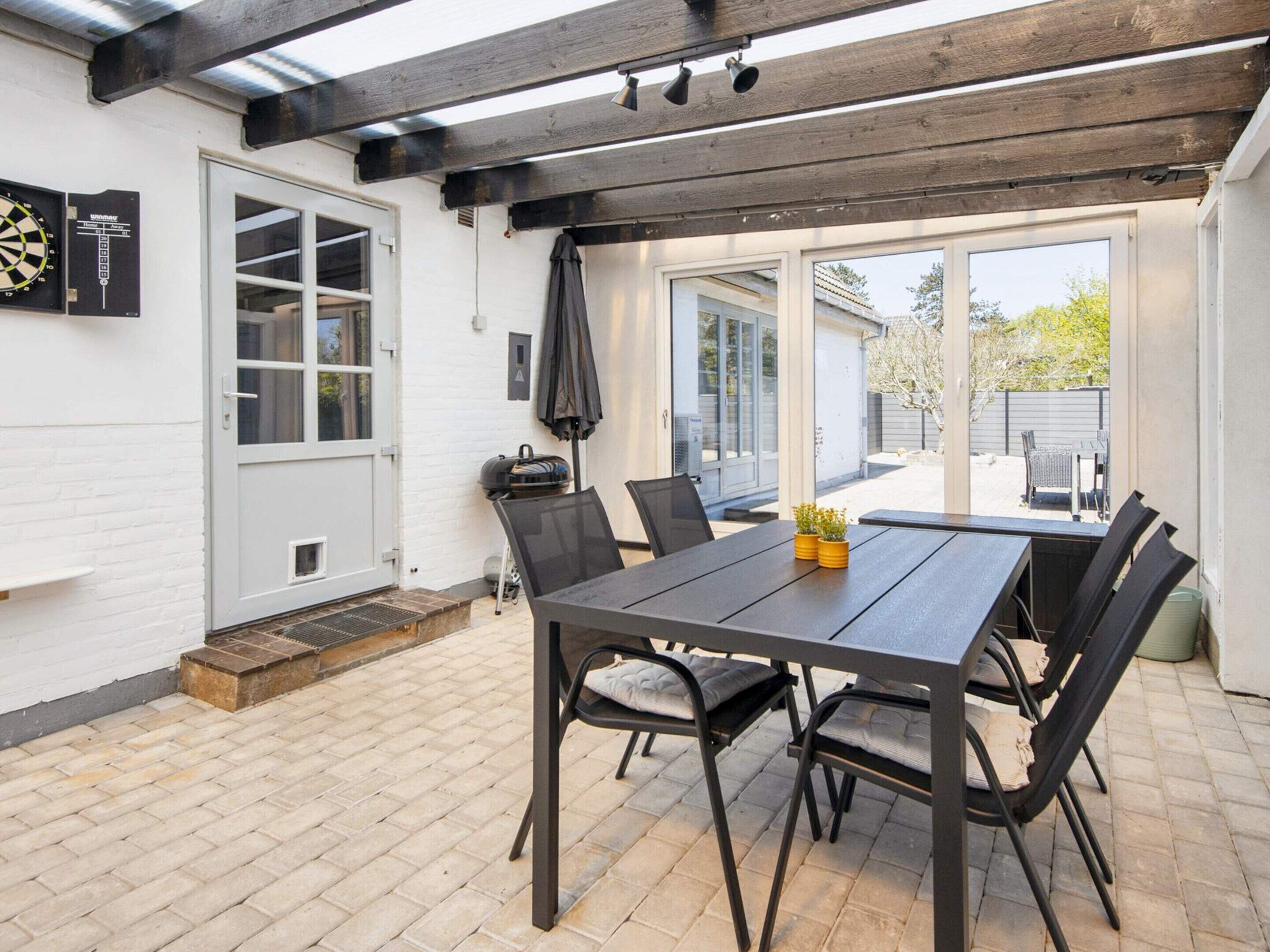 A covered patio with a table and chairs, a door, and a window overlooking a garden.