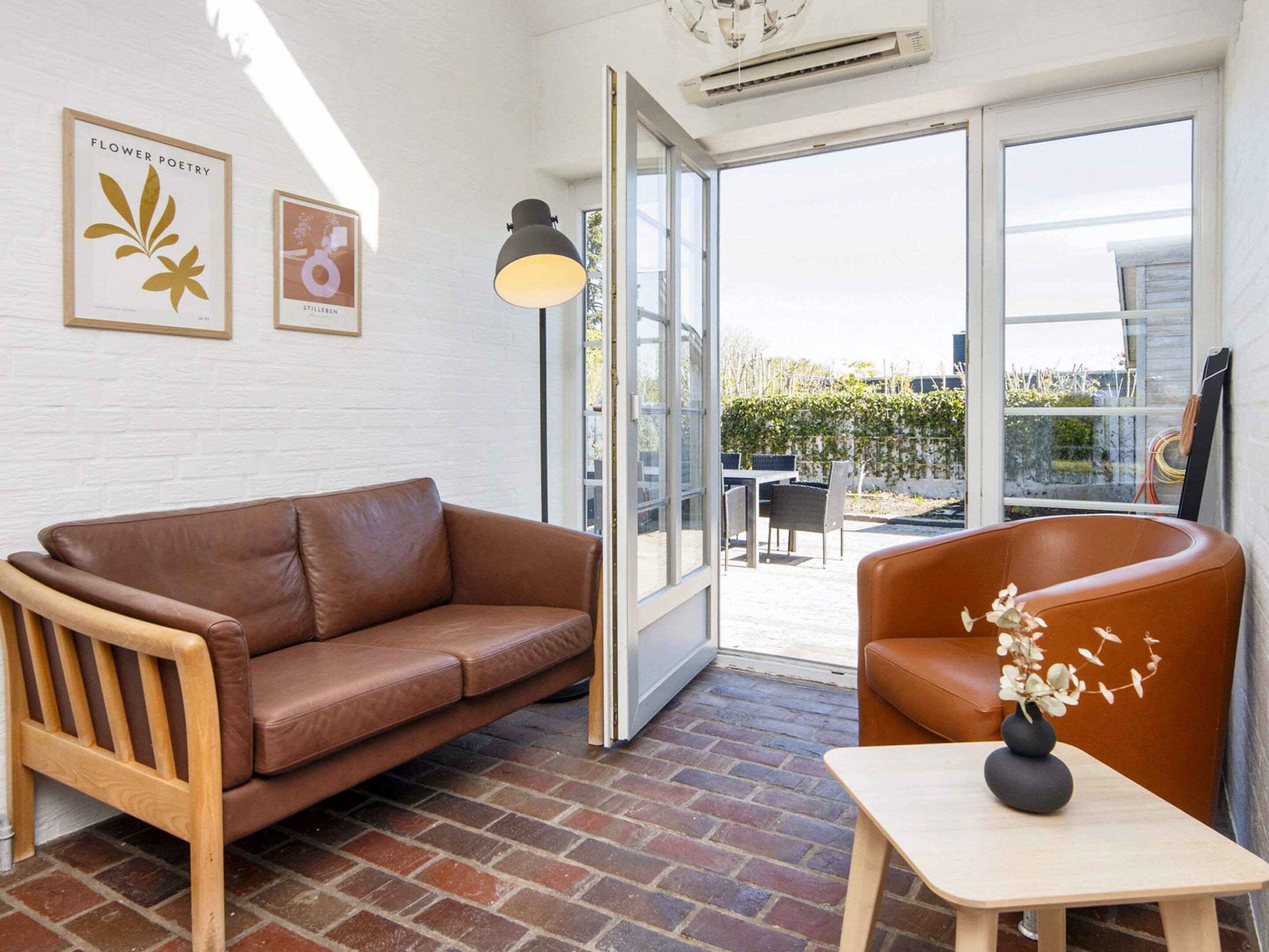 A room with a brown leather sofa, a wooden chair, a small table with a vase, and a large window with a view of the outdoors.