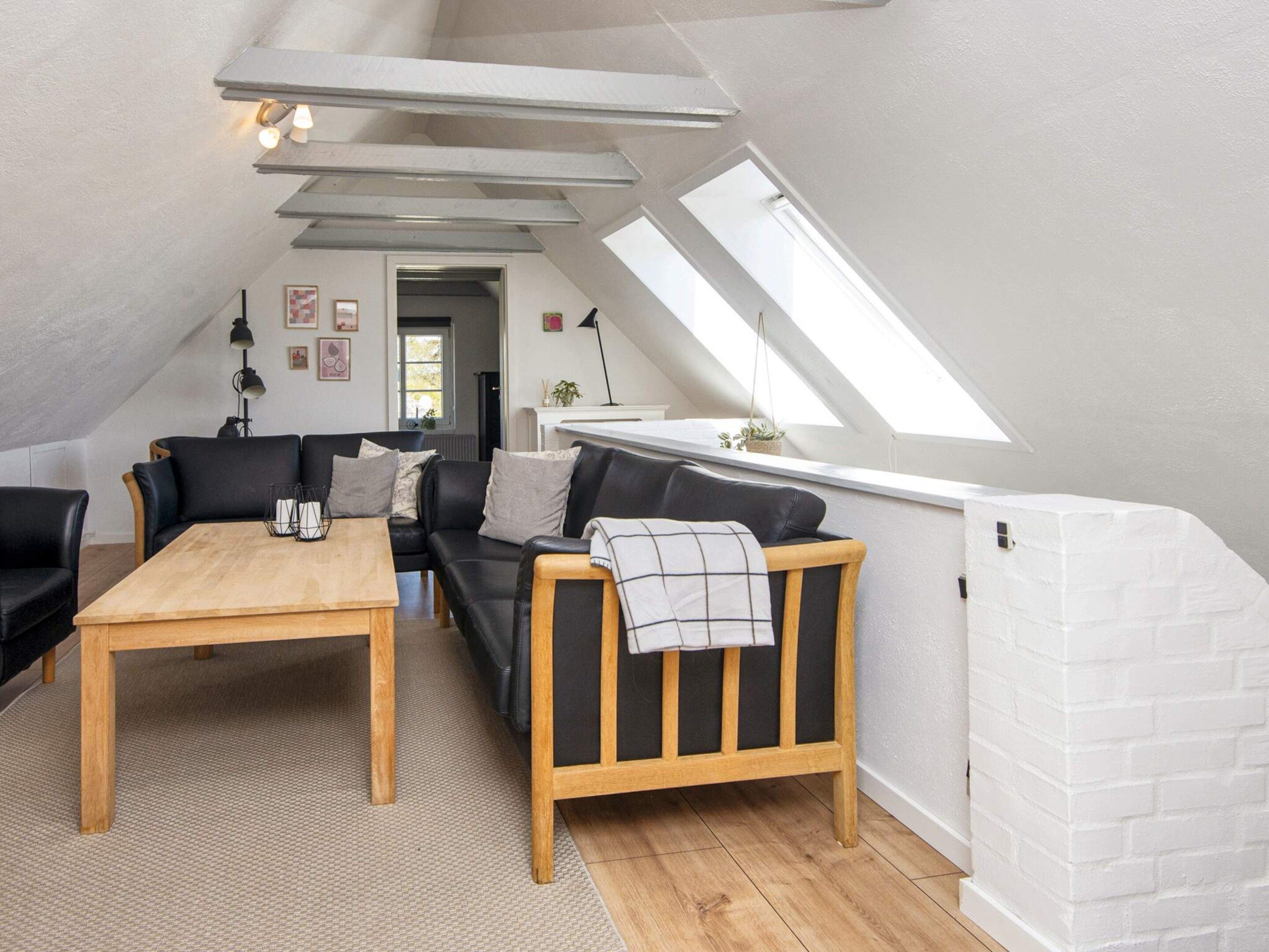 A loft-style room with a sloped ceiling, a wooden table, a black leather sofa, and a white brick accent wall.