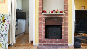 Un salon avec une cheminée en briques, un plancher en bois, un téléviseur et une cuisine à l’arrière-plan.