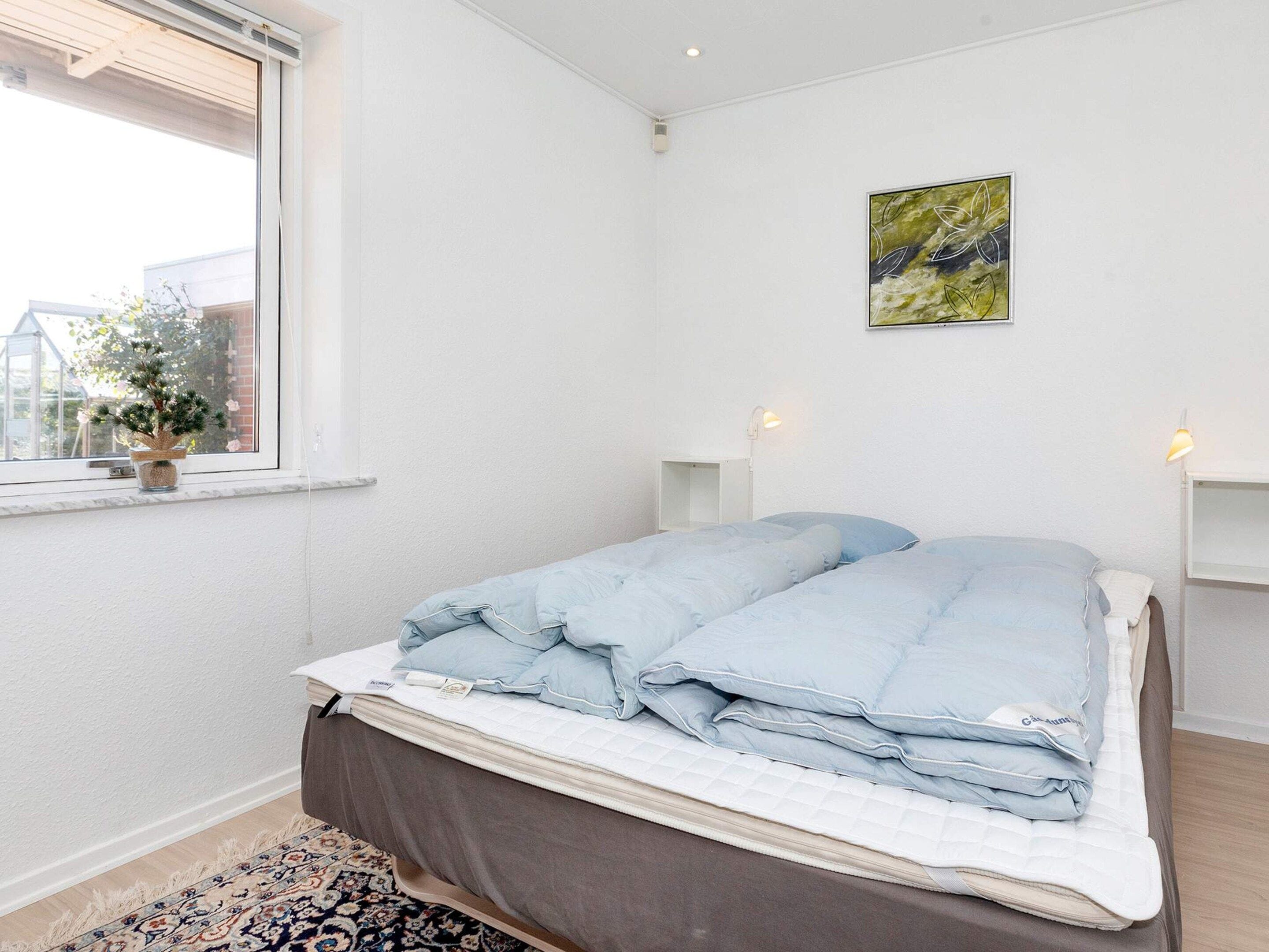 A bedroom with a bed, a window with a plant, a painting on the wall, and a bedside table with a lamp.