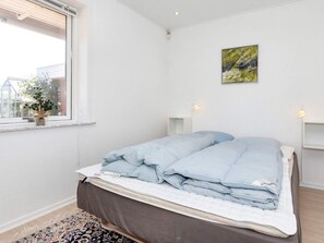 A bedroom with a bed, a window with a plant, a painting on the wall, and a bedside table with a lamp.