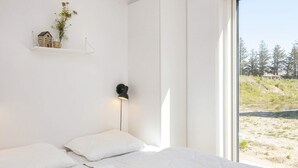 A bedroom with a bed, pillows, a nightstand, a wall-mounted lamp, a shelf with a house model and flowers, and a window with a view of trees and a beach. - 5 Star Holiday Home in Romo (Rømø)