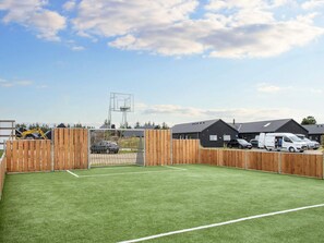 Un terrain de sport clôturé, avec des poteaux de but, entouré de bâtiments et de véhicules stationnés.