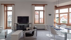 A modern living room with a flat-screen TV, glass dining table, white chairs, and large windows offering a view of buildings. - Stylish Apartment With Balcony/terrace (De Haan)