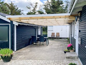 A covered patio area with a table and chairs, a small garden with potted plants, and a view of the surrounding trees. - 6 Person Holiday Home in Lokken (Lokken)
