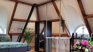 A spacious loft bedroom with a high ceiling, wooden beams, a bed with a floral bedspread, a seating area with a sofa and chairs, and a dining table with a vase of flowers. - Villa in Overloon Near War Museum (Overloon)