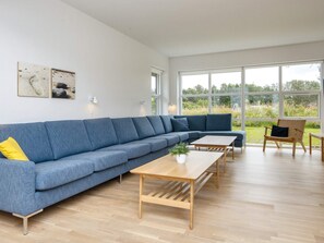 A modern living room with a blue sectional sofa, wooden coffee table, and a view of greenery through large windows. - 14 Person Holiday Park Home in Blavand (Blavand)