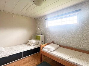 A small, compact room with two single beds, a desk with a lamp, and a window providing natural light.