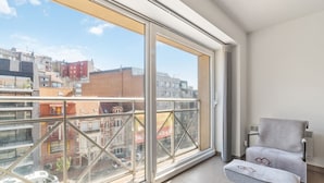 Un apartamento moderno con amplios ventanales de piso a techo, un balcón con barandilla metálica y un sillón gris con una toalla sobre él.