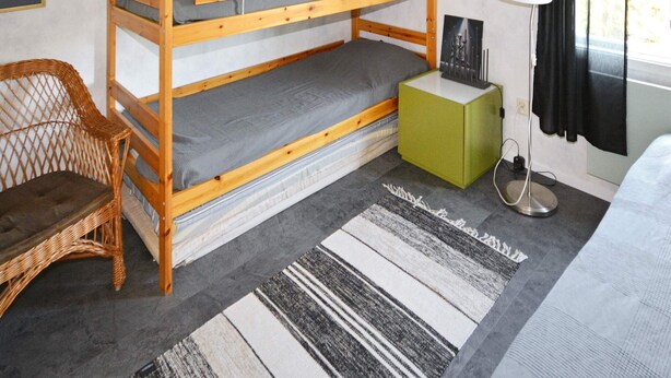 Un lit superposé avec une table de chevet verte, une chaise en osier, une lampe sur pied et un tapis à rayures.