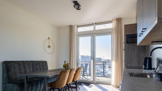 Una sala de estar moderna con un sofá, una mesa de comedor y sillas, un amplio ventanal con vistas a la ciudad, y una zona de cocina con un fregadero y armarios.