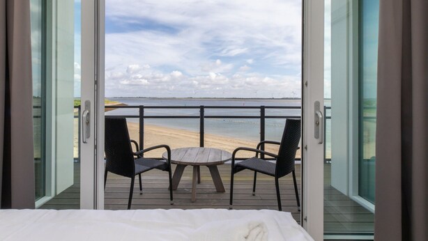 A bedroom with a balcony, a bed, and two chairs facing a beach with wind turbines in the distance. - Seaside Escape, Sint-annaland (Sint-Annaland)