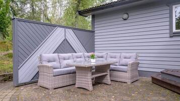 An outdoor seating area with a grey sofa and wicker tables, set against a house with grey siding and a tiled roof.