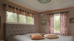 Un dormitorio con cama, dos almohadas, una mesita de noche, ventana con cortinas y un cuadro colgado en la pared.