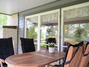 A covered patio with a wooden table and chairs, a potted plant, and a view of greenery through large windows. - 4 Star Holiday Home in Sparreholm (Sparreholm)