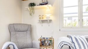 Een gezellige woonkamer met een grijze fauteuil, een klein poufje, een rieten bijzettafeltje met boeken, een lamp en een raam met uitzicht.