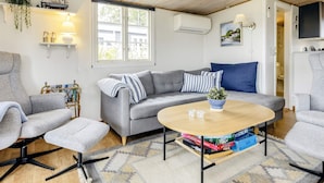 Een moderne woonkamer met een grijze bank, een ronde salontafel en een grijze fauteuil. Er is een raam met uitzicht op gebouwen, een wandairconditioner en een klein plankje met decoratieve objecten.