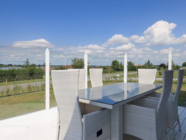 Spisebord med glasplade og hvide fletstole, placeret på en terrasse med udsigt over en sø og fjerne bygninger.