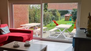 A modern living room with a red sofa, a wooden coffee table, and a sliding glass door leading to an outdoor patio with picnic tables and a small wooden playhouse. - Canal-side Family Retreat (Nieuwpoort)