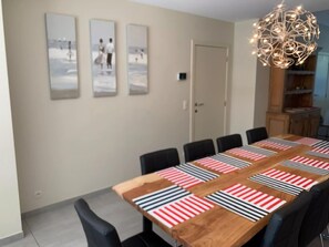 A dining area with a long wooden table, black chairs, and striped table mats. There are three wall-mounted pictures depicting beach scenes. A modern chandelier hangs above the table. - Canal-side Family Retreat (Nieuwpoort)
