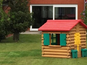 A small wooden playhouse with a red roof and green shutters, situated in a grassy area with a tree and a house in the background. - Canal-side Family Retreat (Nieuwpoort)