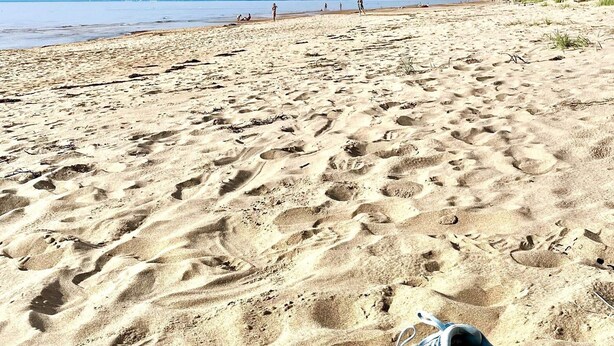 Schoenen in het zand op een strand, met voetafdrukken, een heldere lucht en een rustige zee op de achtergrond.