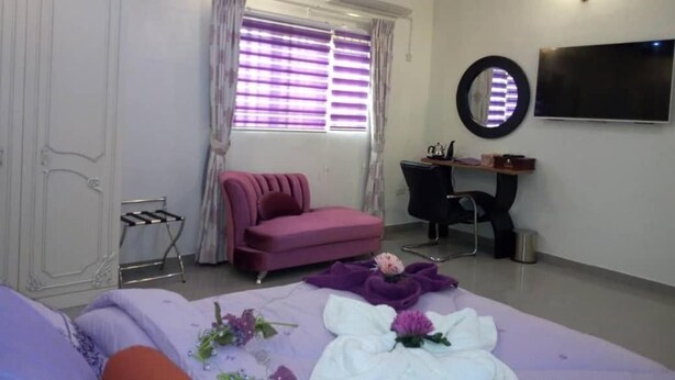A bedroom with a bed, a desk, a chair, a window with curtains, and a TV on the wall. - Lavender Spa and Suites (Enugu)
