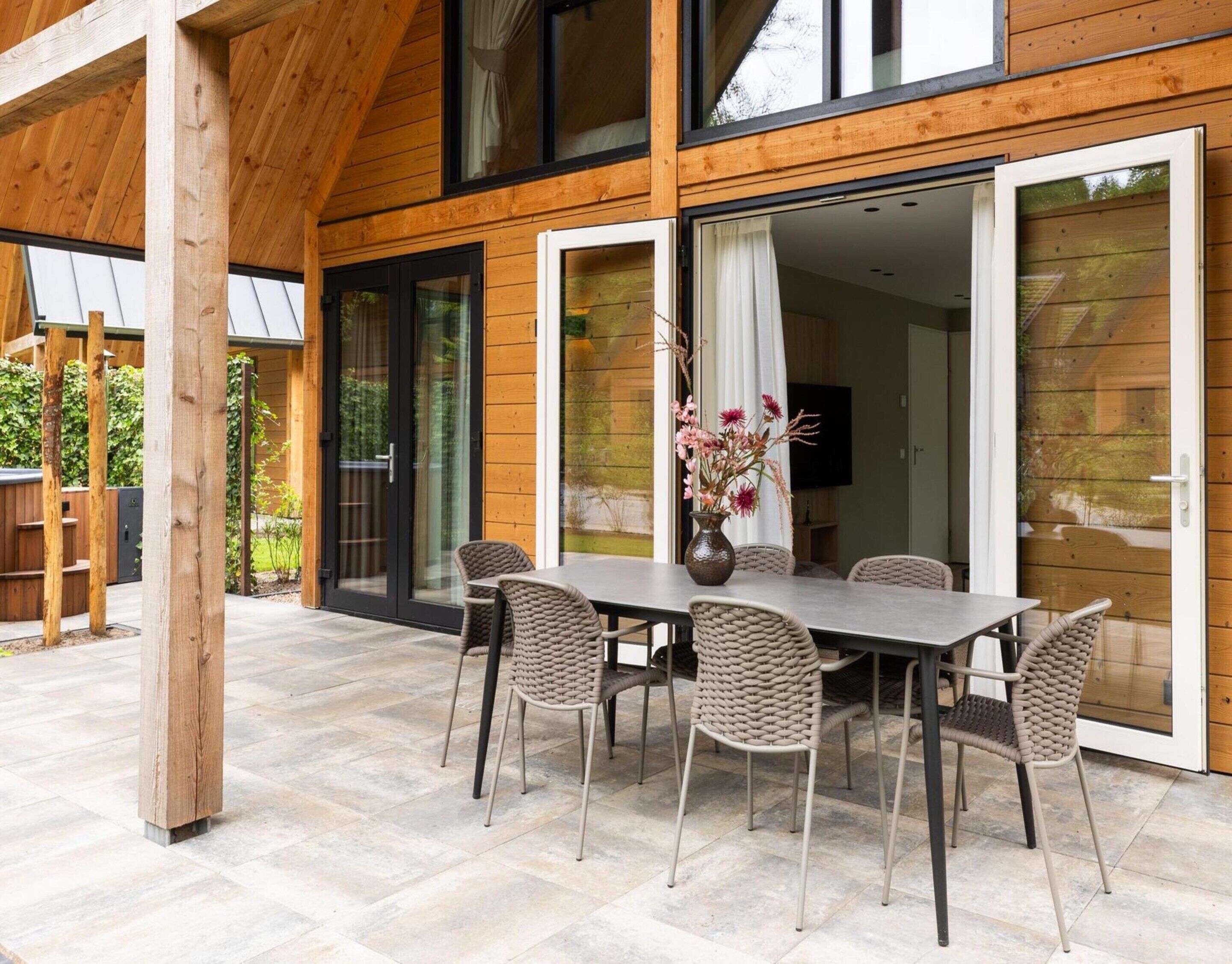 Fachada de madera con un patio cubierto, una mesa de comedor con sillas y un jarrón con flores.