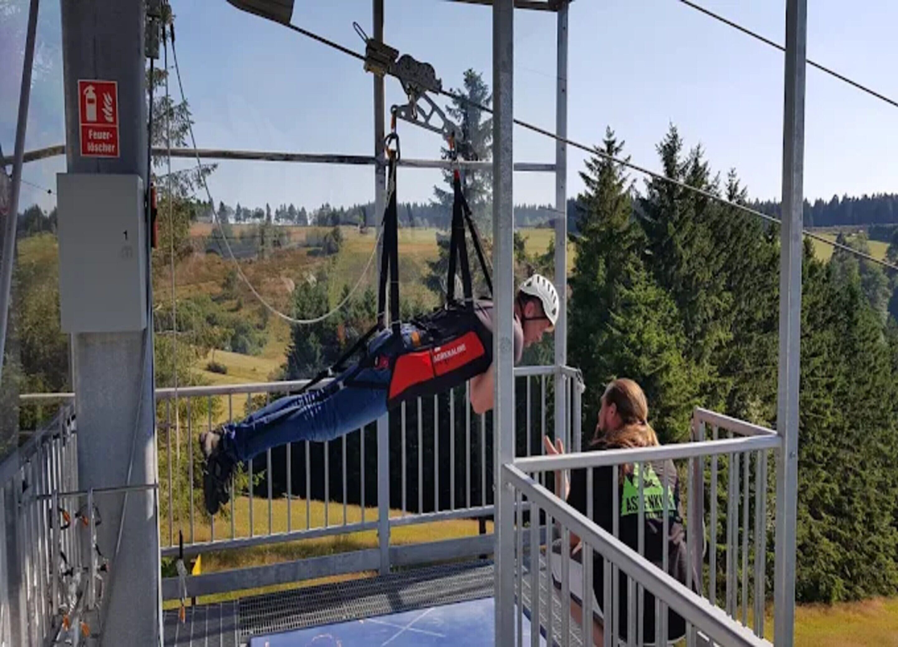 A cable car with a person in a harness, a railing, and a view of a forested landscape.