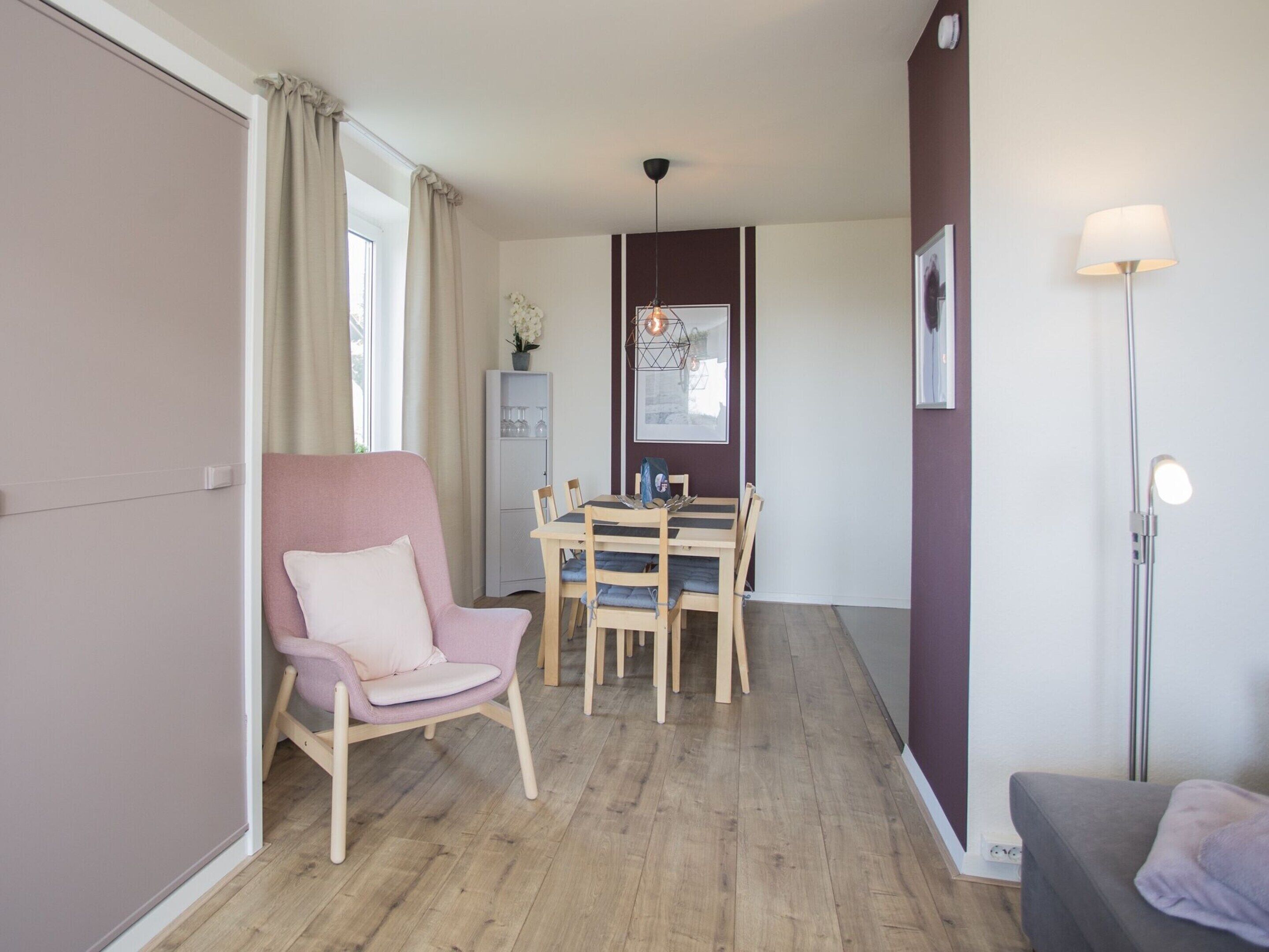 A cozy room with a wooden floor, a pink armchair, a dining table with chairs, a cabinet, and a floor lamp.