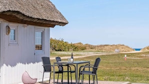 Una cabaña con techo de paja y una mesa con sillas en un patio empedrado, con vistas a un área verde y al mar.