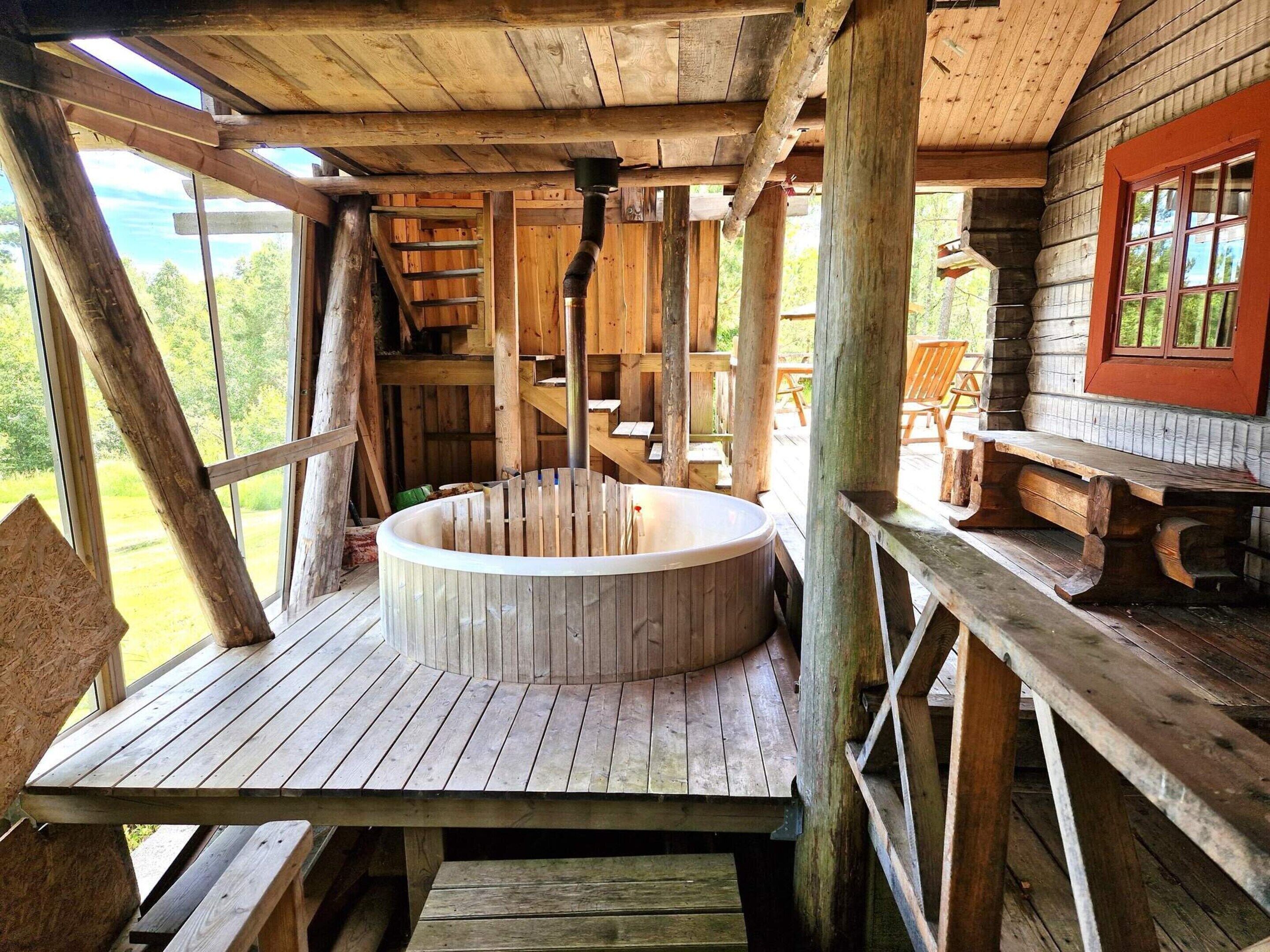 Una bañera de madera en una cabaña rústica con terraza de madera y ventana con vistas a la vegetación.