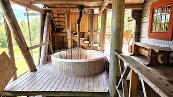 Una bañera de madera en una cabaña rústica con terraza de madera y ventana con vistas a la vegetación.