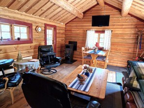 Interior acolhedor de cabana com teto de madeira, lareira, mesa de jantar com cadeiras, poltrona de couro e televisão na parede.