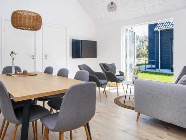 A modern interior with a wooden dining table, grey upholstered chairs, a flat-screen TV, and a view of a blue building through glass doors.
