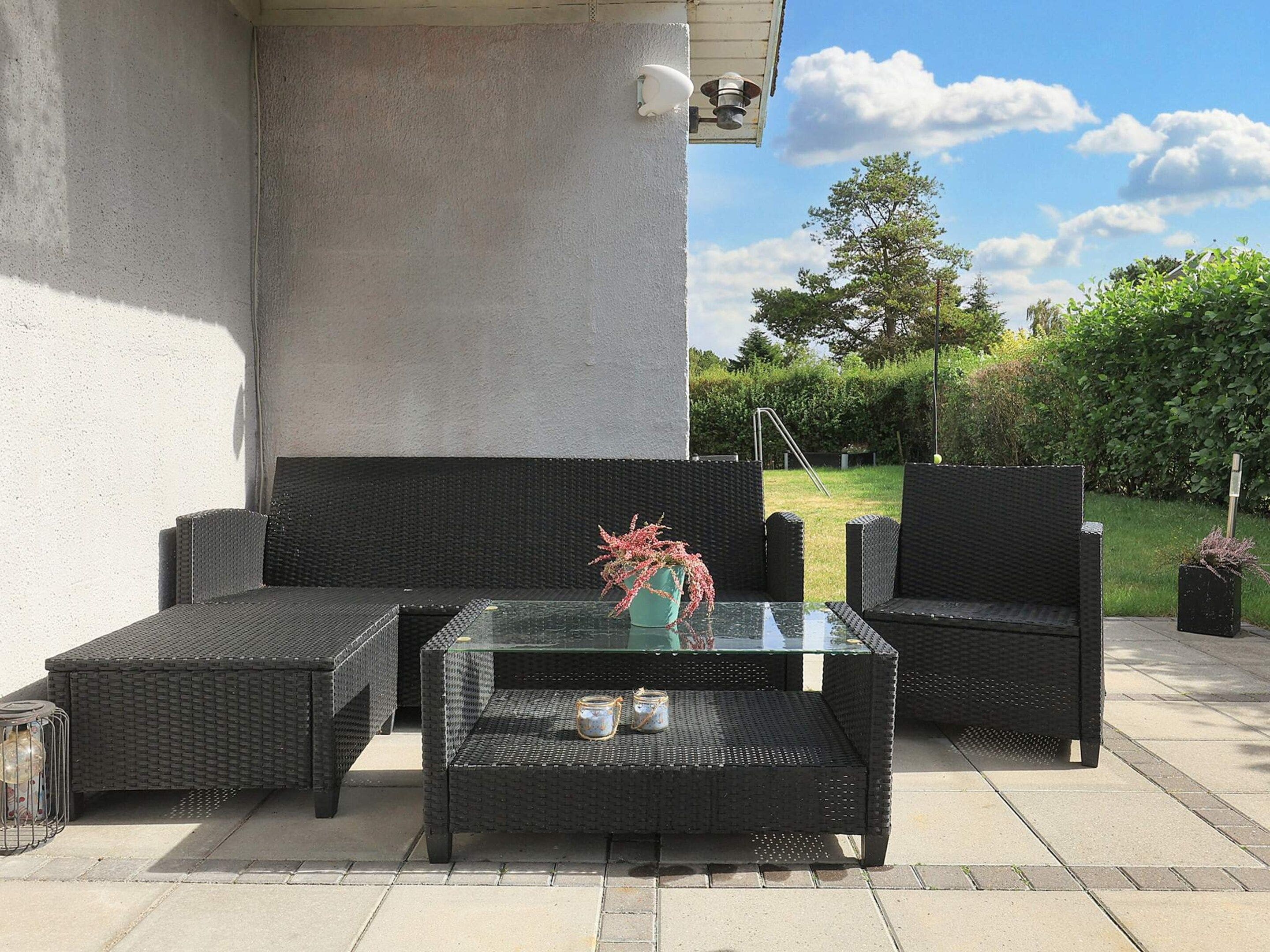 Un espace extérieur aménagé avec des meubles en osier et une table à plateau de verre, situé devant un bâtiment doté d’un mur en béton, le tout sous un ciel dégagé.