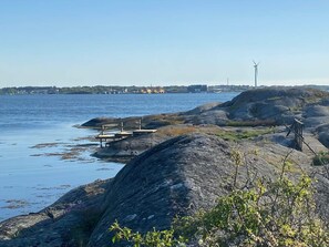 En klippefyldt kystlinje med en træbro, der strækker sig ud i rolige farvande, en vindmølle i det fjerne, og en klar himmel.