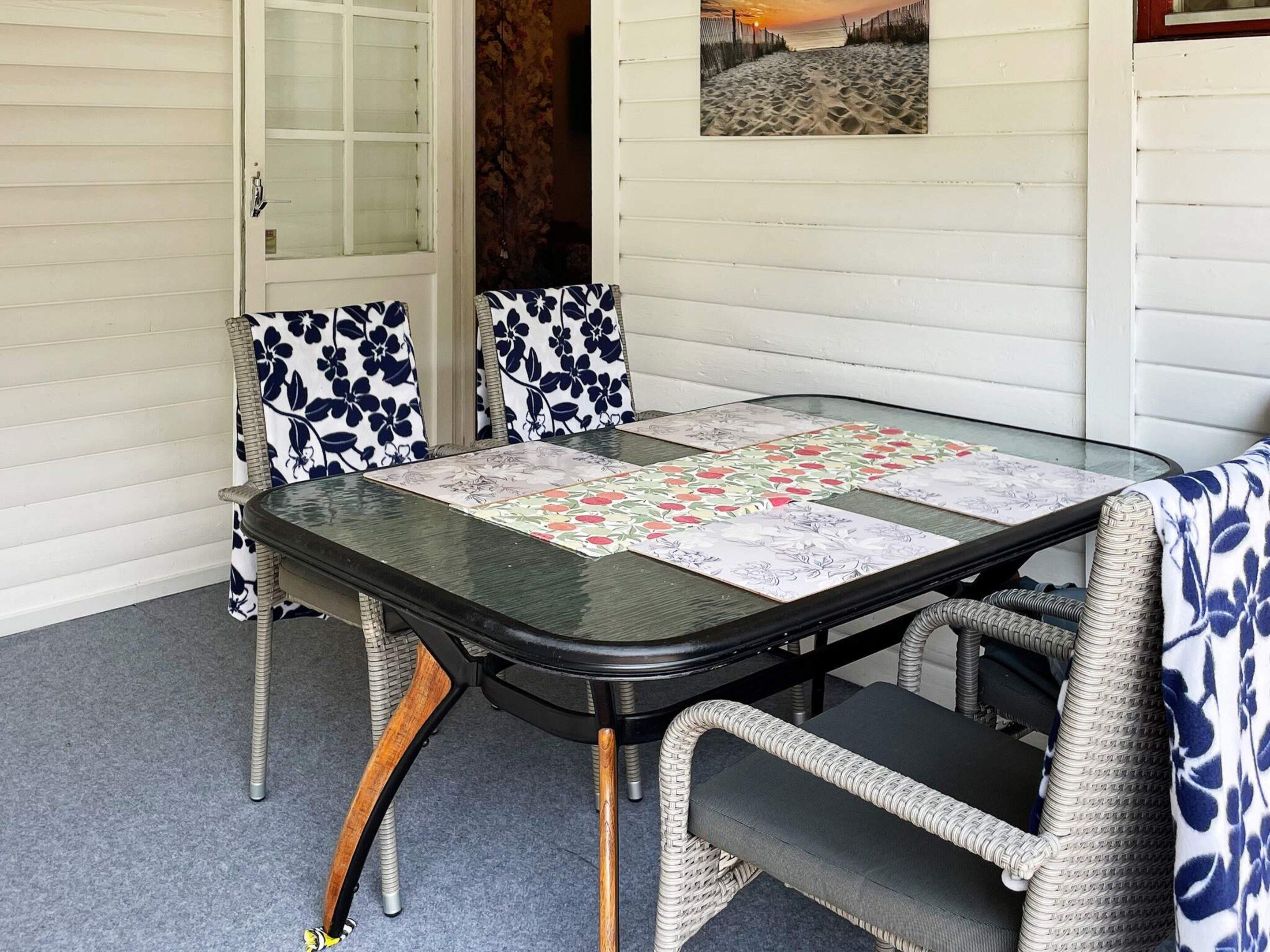 Mesa de jantar com tampo de vidro e um jogo americano floral, rodeada por cadeiras de vime com almofadas florais em azul e branco.