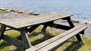 Une table de pique-nique en bois, située au bord d’un lac, avec des galets et de l’herbe, et une île visible au loin.